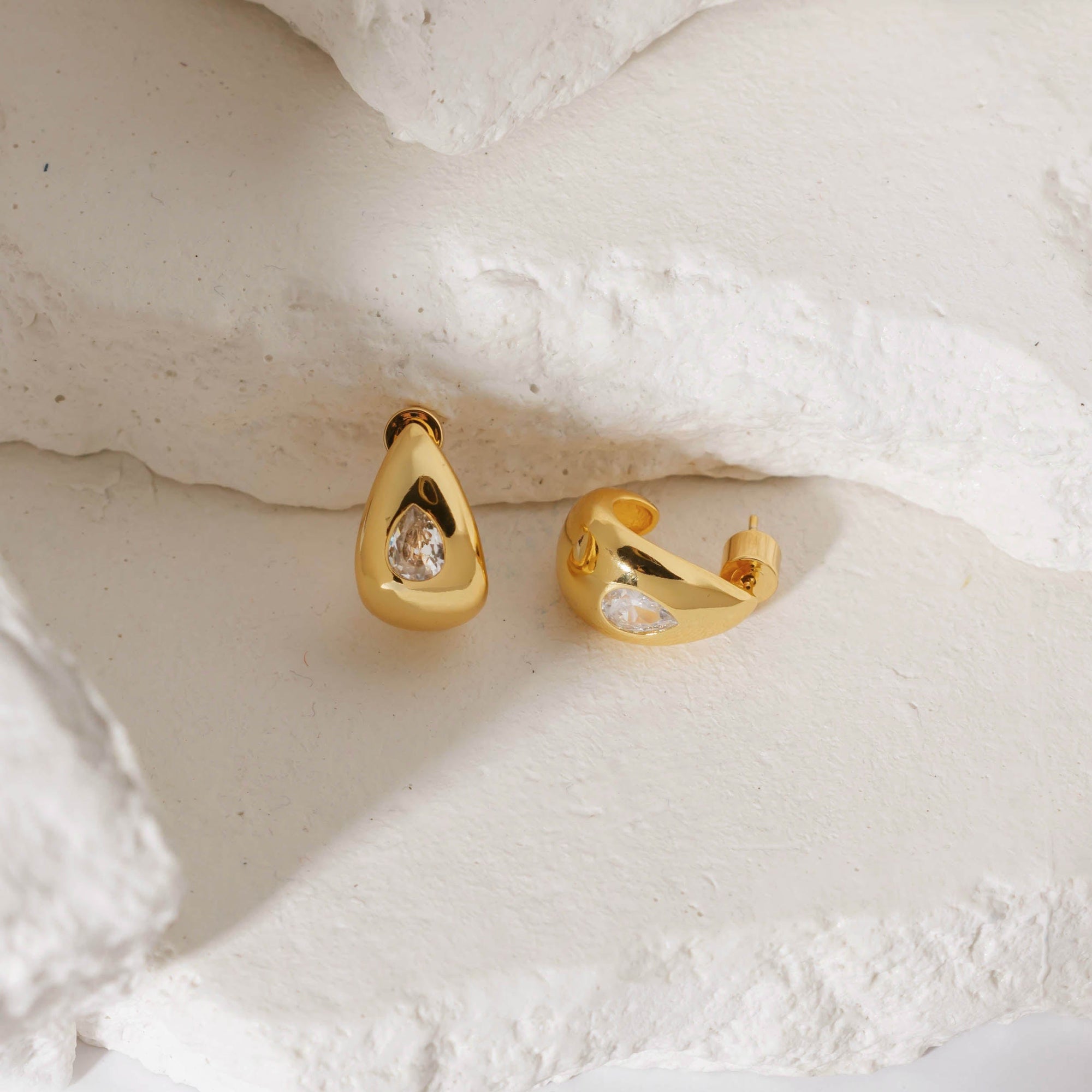 A pair of Amadora Hoop earrings with crystal accents on white stone slabs, one earring standing tall on its curve as the other lay on its side by The Perfect Hoop.
