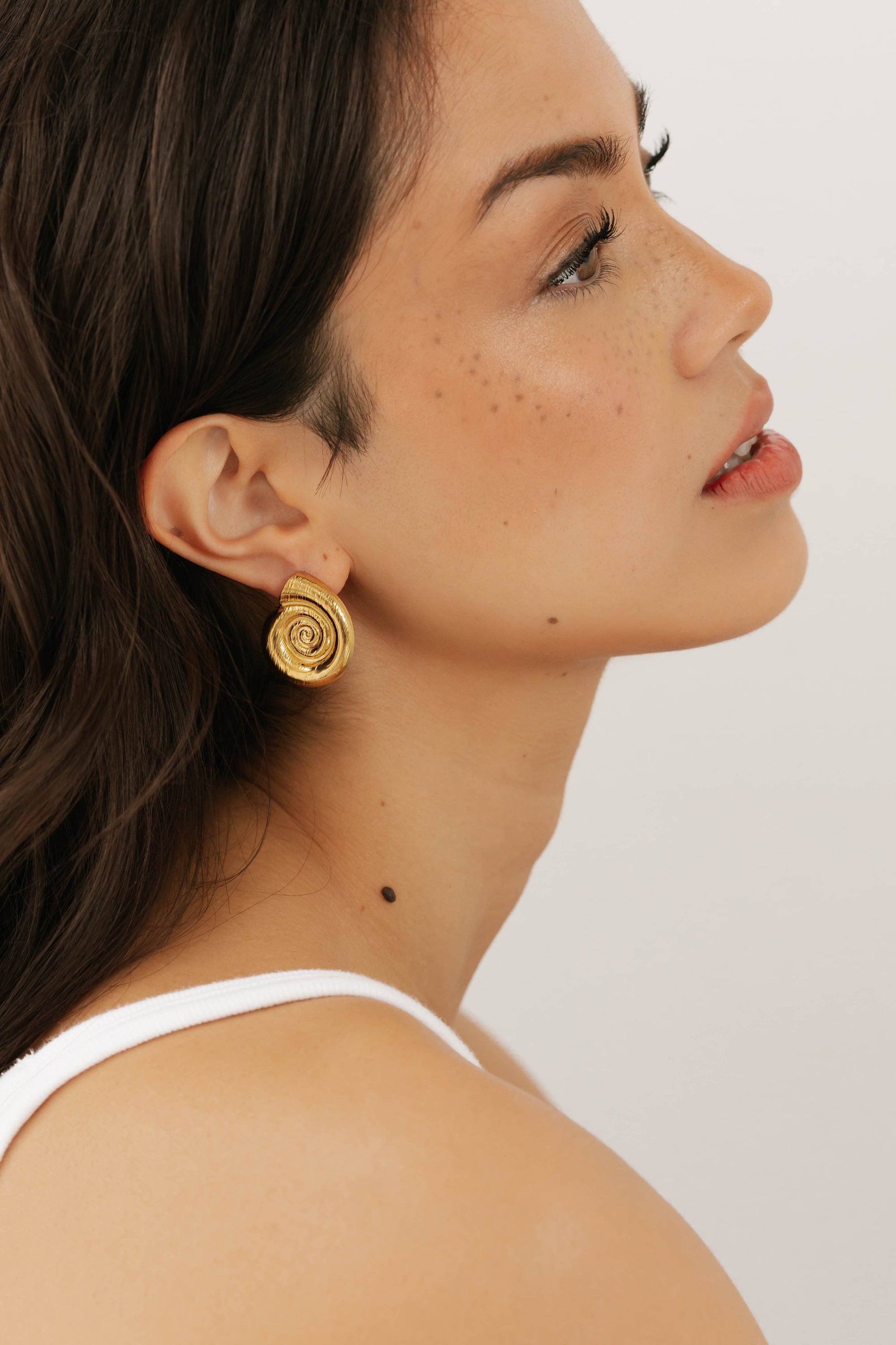 Model wearing the gold By The Sea Earring with a subtle smile, styled against a clean white background.  By The Perfect Hoop.