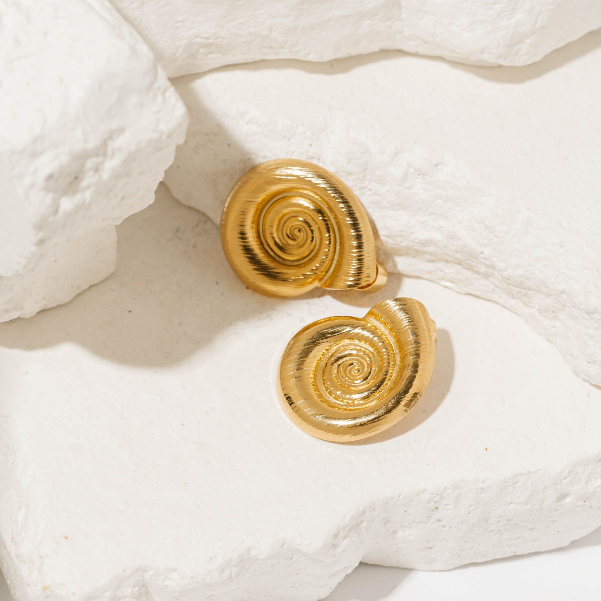 A pair of gold, spiral shell-inspired By The Sea Earrings displayed on textured white stones.  By The Perfect Hoop.